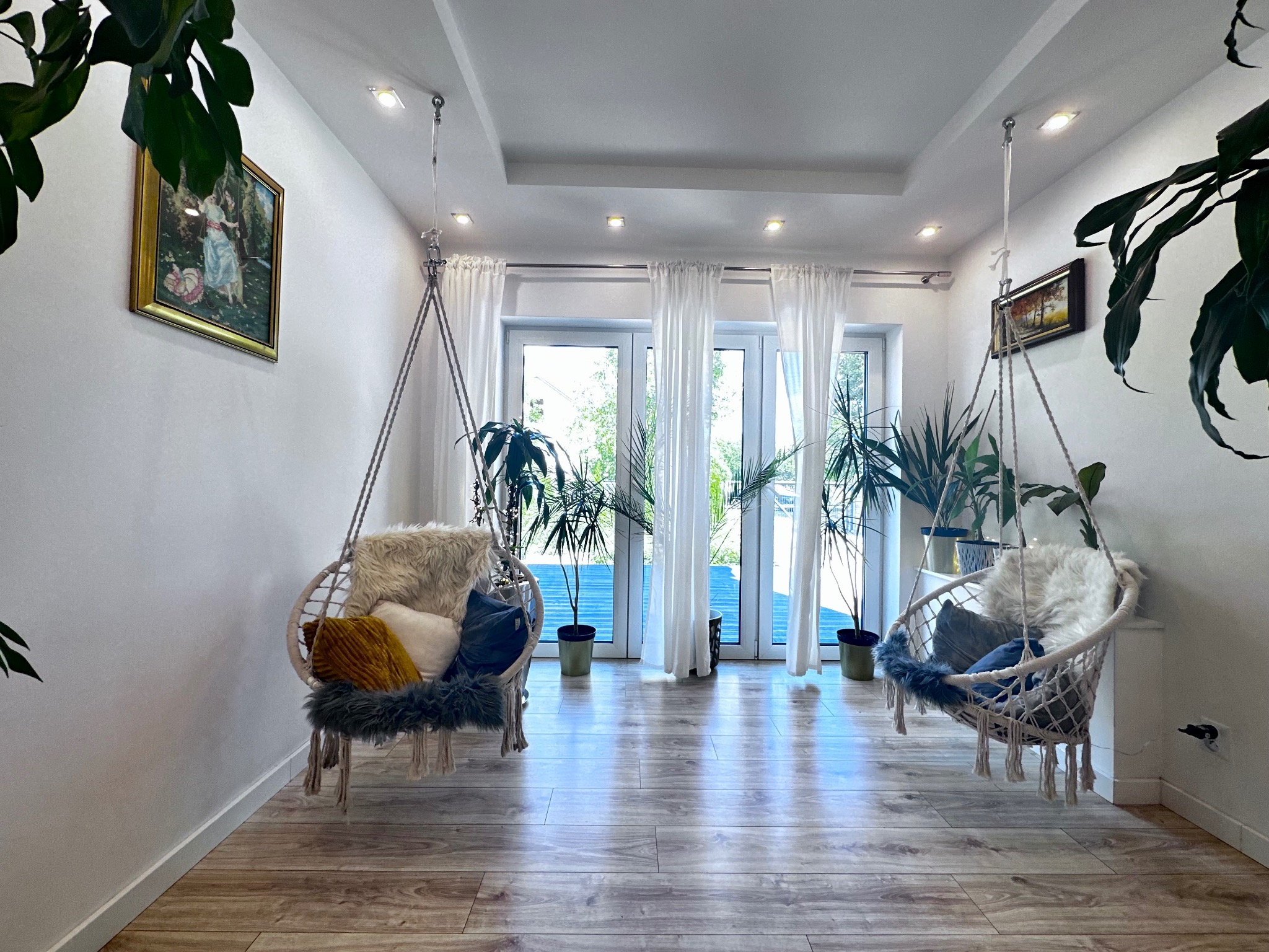 Jasne wnętrze z dwoma wiszącymi fotelami typu kokon, dekorowane poduszkami i futrzanymi narzutami, z parkietem i roślinami doniczkowymi, widok na taras przez przeszklone drzwi balkonowe.