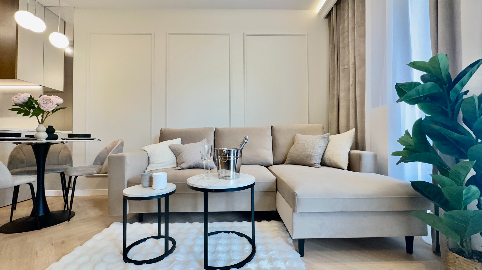 Beżowy narożnik z poduszkami, stoliki kawowe z szampanem, kwiaty i roślina doniczkowa w jasnym salonie z panelami ściennymi i drewnianą podłogą.