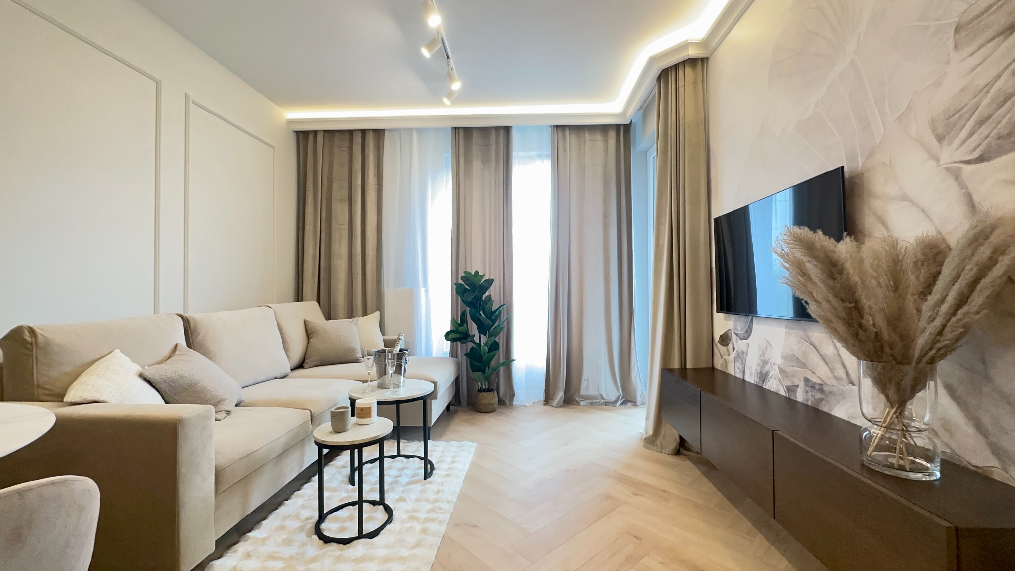 Jasne, przestronne wnętrze salonu z beżową sofą, stolikami kawowymi i telewizorem na ścianie z motywem roślinnym. Parkiet w jodełkę, dekoracyjne zasłony i subtelne oświetlenie.