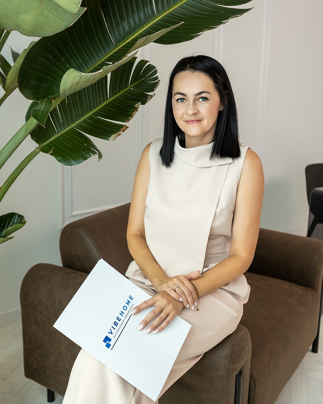 Profesjonalna agentka nieruchomości VibeHome siedzi na fotelu z dokumentami, w tle roślina i jasne ściany. Uśmiechnięta, elegancka, w beżowym stroju.