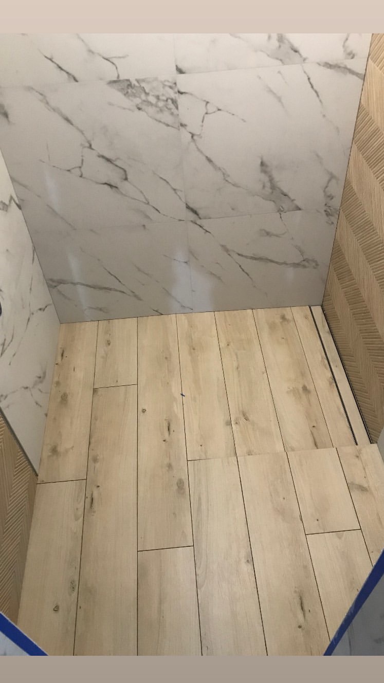 Wykończona kabina prysznicowa z drewnopodobną podłogą i marmurowymi płytkami ściennymi, widoczny odpływ liniowy i dekoracyjna ściana z wzorem jodełki.