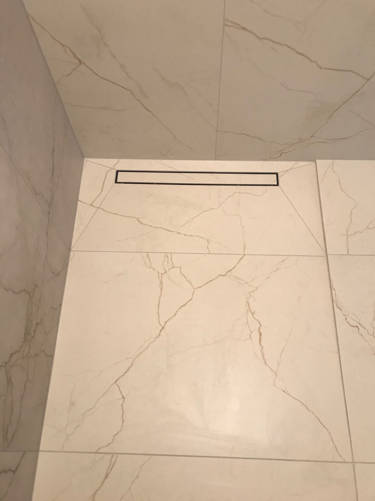 Płytki podłogowe i ścienne z jasnego marmuru z ciemnymi żyłkami, czarny odpływ liniowy w rogu kabiny prysznicowej.