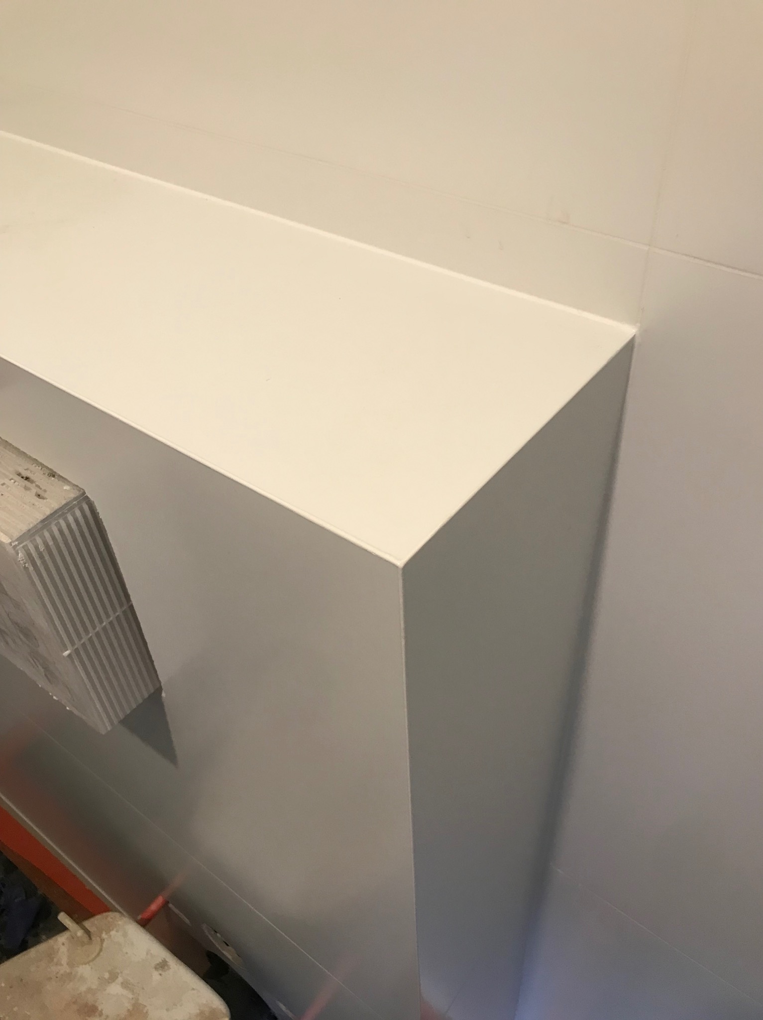 Biała, gładka zabudowa z płyty kartonowo-gipsowej w rogu pomieszczenia, widoczne łączenia płyt i fragment brudnego wiadra z zaprawą w dolnym rogu.