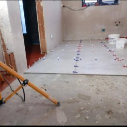 F.R.U - Bart.Ew - Wnętrze w trakcie układania płytek podłogowych, widoczne białe płytki z niebieskimi i czerwonymi klipsami poziomującymi, wiaderka z zaprawą, lampa budowlana i fragment osoby pracującej w tle.