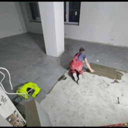 F.R.U - Bart.Ew - Pracownik w czerwonym kombinezonie układa szare płytki podłogowe w pomieszczeniu z białymi ścianami i filarem, widoczna grzebieniowa paca i poziomica.