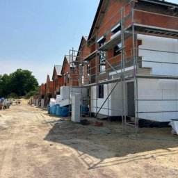 F.R.U - Bart.Ew - Rząd domów w trakcie budowy, widoczne rusztowania, ocieplenie styropianem i cegła, teren budowy z workami i materiałami.