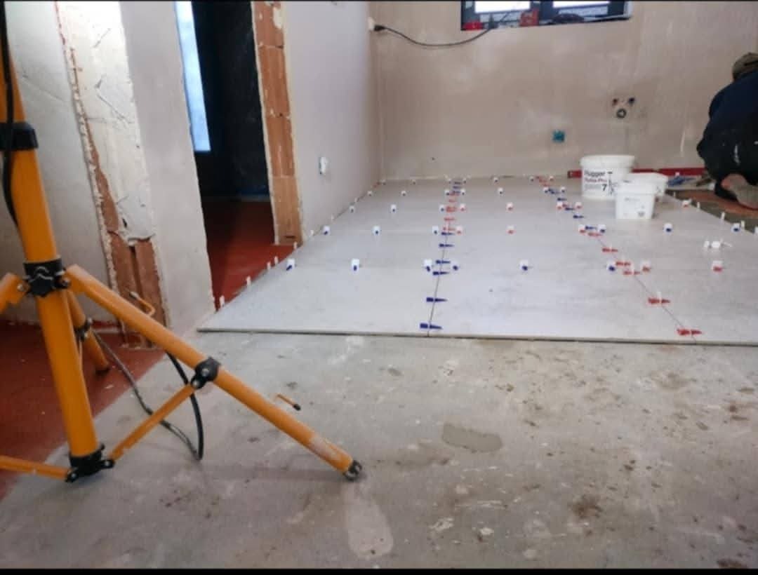 Wnętrze w trakcie układania płytek podłogowych, widoczne białe płytki z niebieskimi i czerwonymi klipsami poziomującymi, wiaderka z zaprawą, lampa budowlana i fragment osoby pracującej w tle.