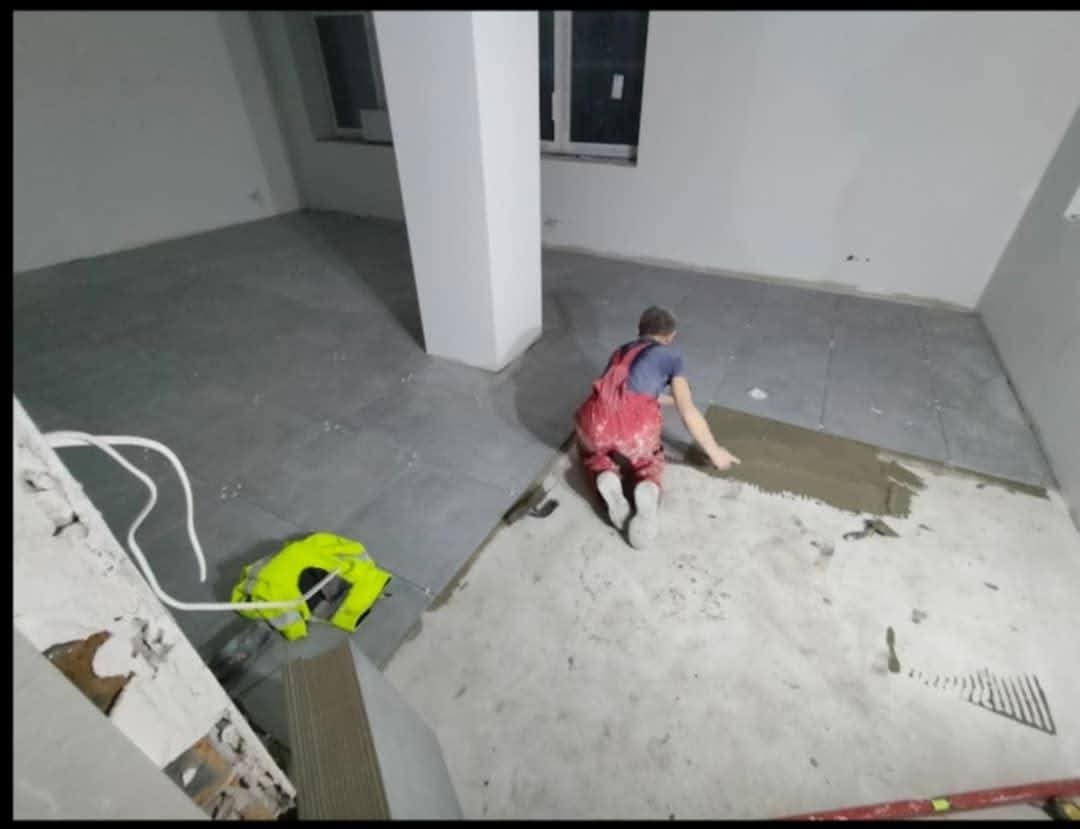 Pracownik w czerwonym kombinezonie układa szare płytki podłogowe w pomieszczeniu z białymi ścianami i filarem, widoczna grzebieniowa paca i poziomica.