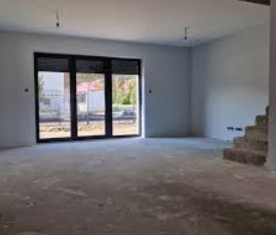 Surowe wnętrze domu w trakcie wykańczania, widoczne betonowe schody, surowa podłoga i duże okno balkonowe z czarnymi ramami, przez które widać zieleń.