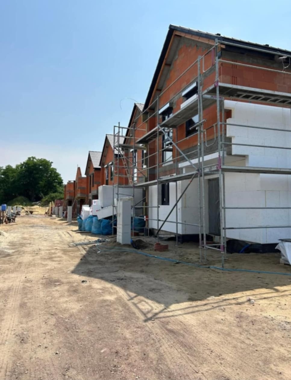 Rząd domów w trakcie budowy, widoczne rusztowania, ocieplenie styropianem i cegła, teren budowy z workami i materiałami.