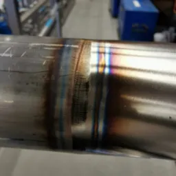 Szlifowany spaw na metalowej rurze z widocznymi śladami obróbki i tęczowymi przebarwieniami od temperatury, w tle warsztat.