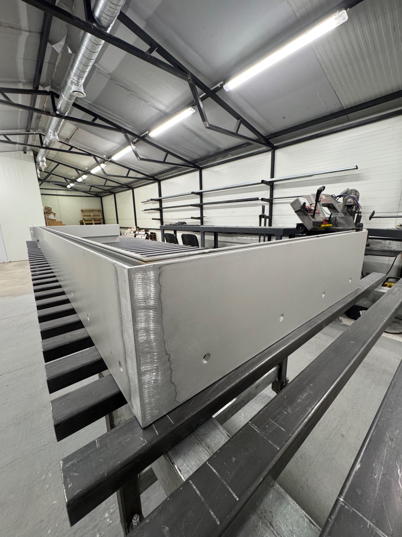 Szara, metalowa rama w warsztacie, widoczna spawana konstrukcja, otwory montażowe i maszyna do cięcia metalu w tle. Surowy, industrialny charakter.