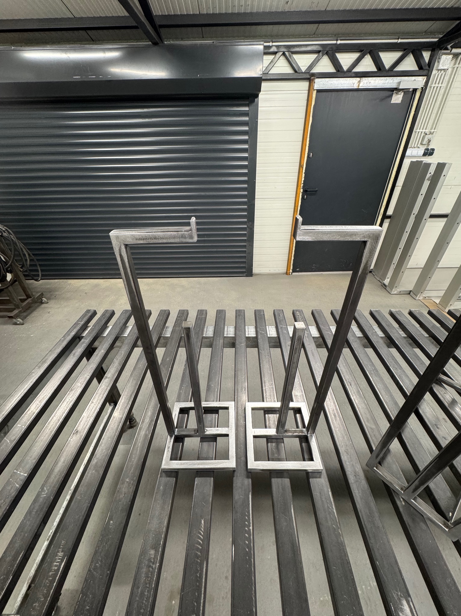 Metalowe elementy konstrukcyjne, prawdopodobnie do bramy, ułożone na metalowych profilach w warsztacie. W tle brama rolowana i drzwi.