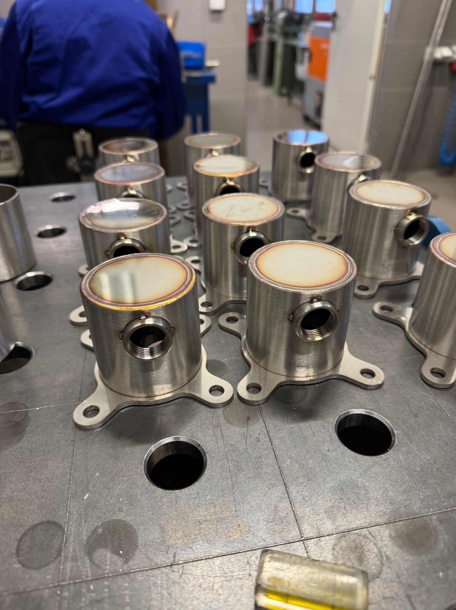 Szereg metalowych, cylindrycznych elementów z podstawami na perforowanym stole spawalniczym. Widoczne ślady spawania na górnych krawędziach. W tle osoba w niebieskim uniformie.
