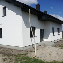 MAR-BUD Usługi budowlano- wykończeniowe - Nowoczesny dom z białą elewacją i ciemną stolarką okienną, widoczny wąż wentylacyjny podczas prac wykończeniowych, na tle błękitnego nieba z chmurami.