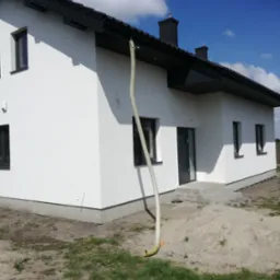 Nowoczesny dom z białą elewacją i ciemną stolarką okienną, widoczny wąż wentylacyjny podczas prac wykończeniowych, na tle błękitnego nieba z chmurami.