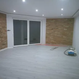 Pokój po remoncie: ściany z dekoracyjnej cegły, jasna podłoga, białe ściany i sufit z oświetleniem punktowym, widoczne narzędzia i odkurzacz.