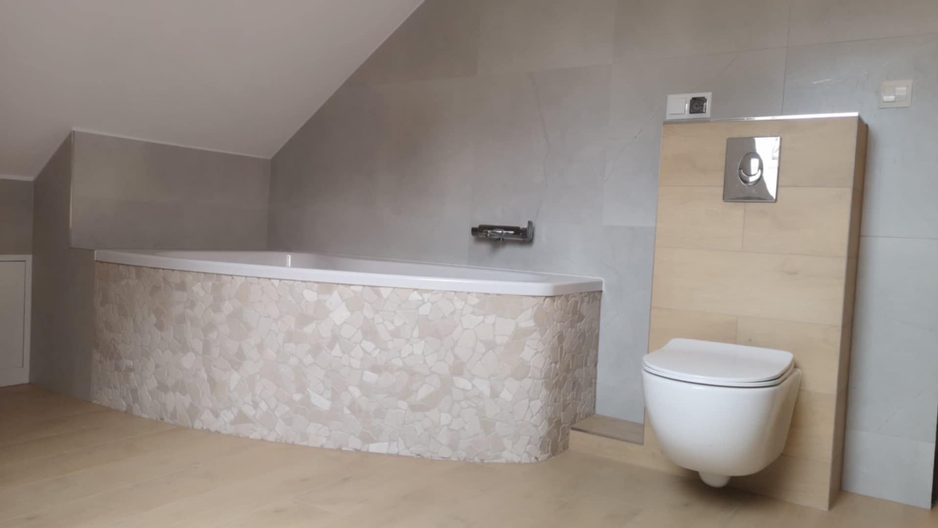 Nowoczesna łazienka na poddaszu: wanna obudowana mozaiką z jasnych kamieni, podwieszana toaleta z drewnianym panelem, szare płytki na ścianach i jasna drewniana podłoga.