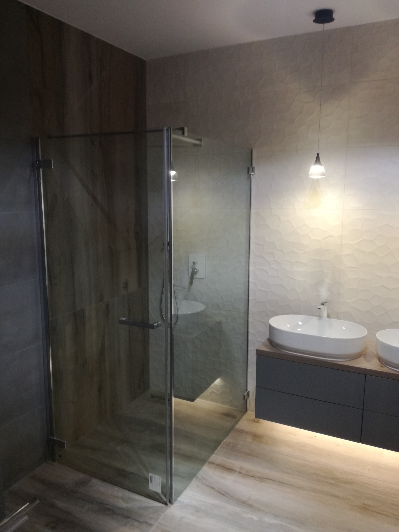 Nowoczesna łazienka z kabiną prysznicową ze szklanymi drzwiami, dwiema owalnymi umywalkami nablatowymi na szarej szafce i podświetleniem LED pod szafką oraz dekoracyjną ścianą z wzorem 3D.