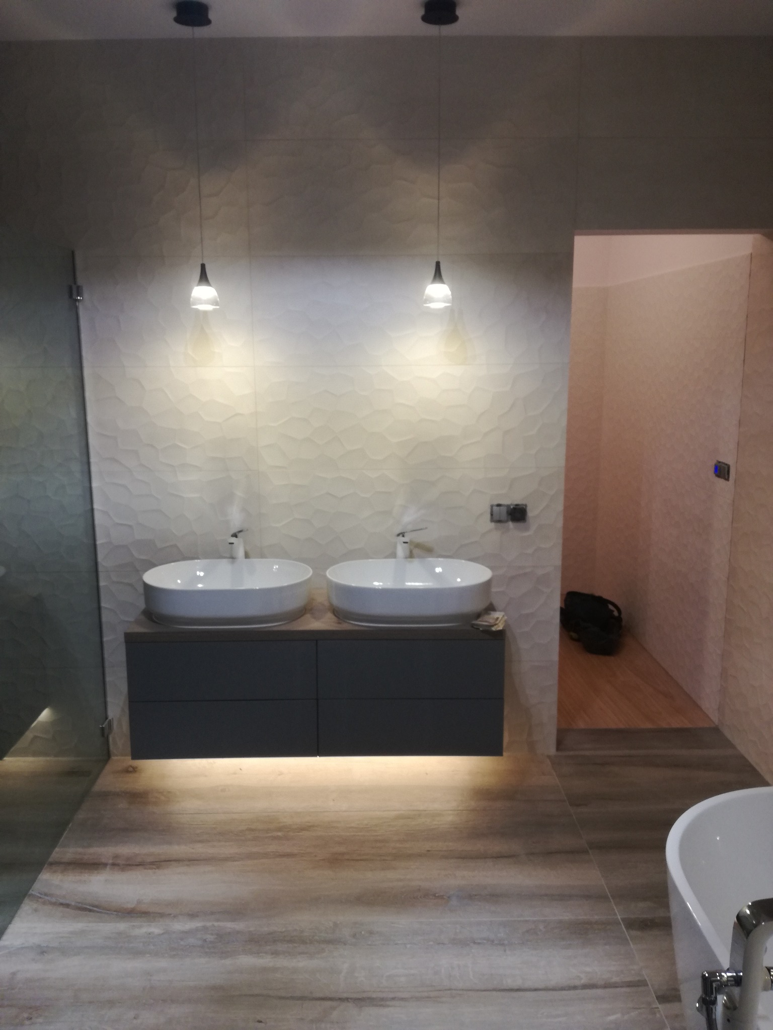 Nowoczesna łazienka z dwoma umywalkami nablatowymi, szarą szafką pod umywalką, podświetleniem LED, teksturowaną ścianą w tle i podłogą imitującą drewno.