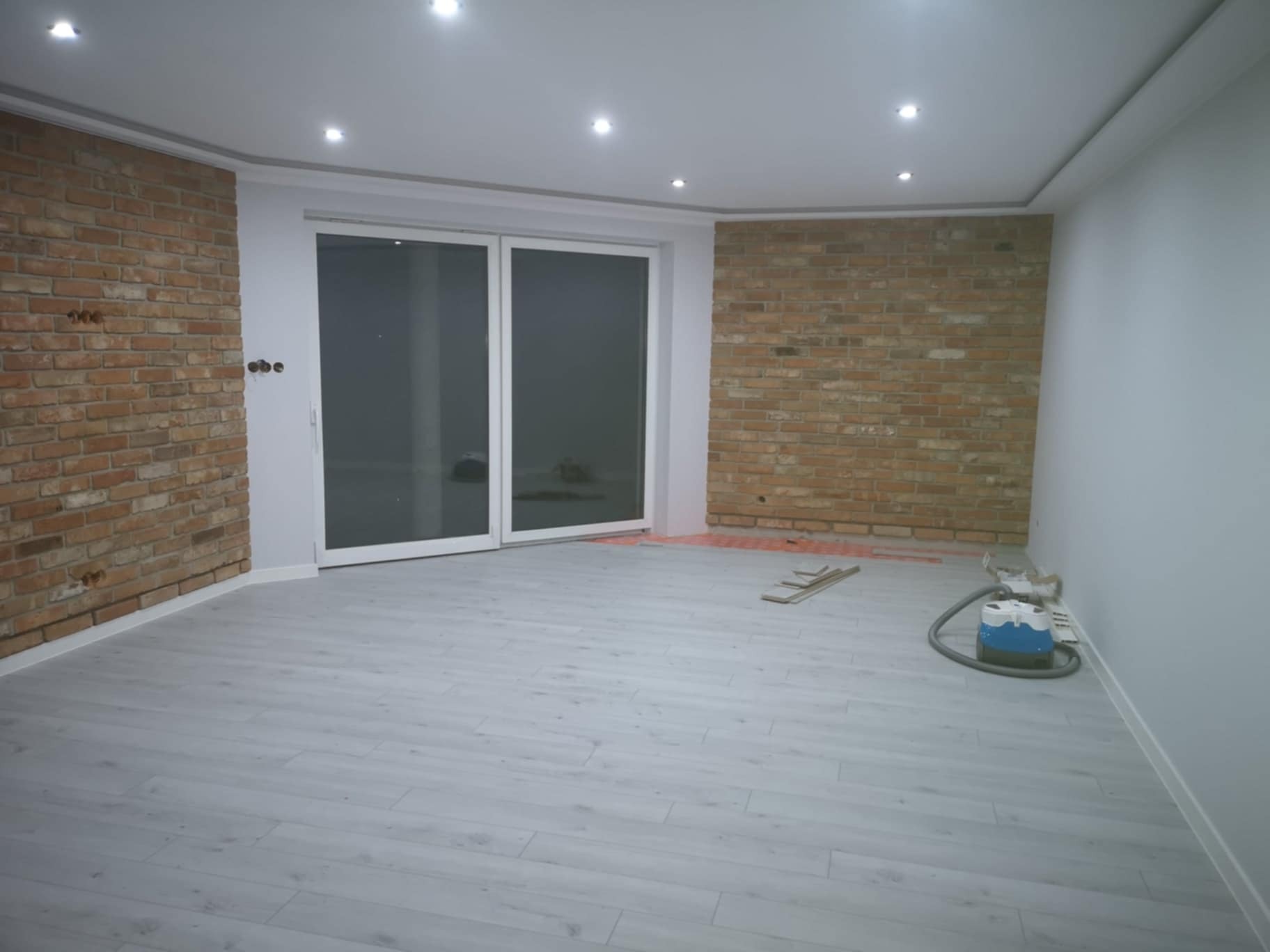 Pokój po remoncie: ściany z dekoracyjnej cegły, jasna podłoga, białe ściany i sufit z oświetleniem punktowym, widoczne narzędzia i odkurzacz.