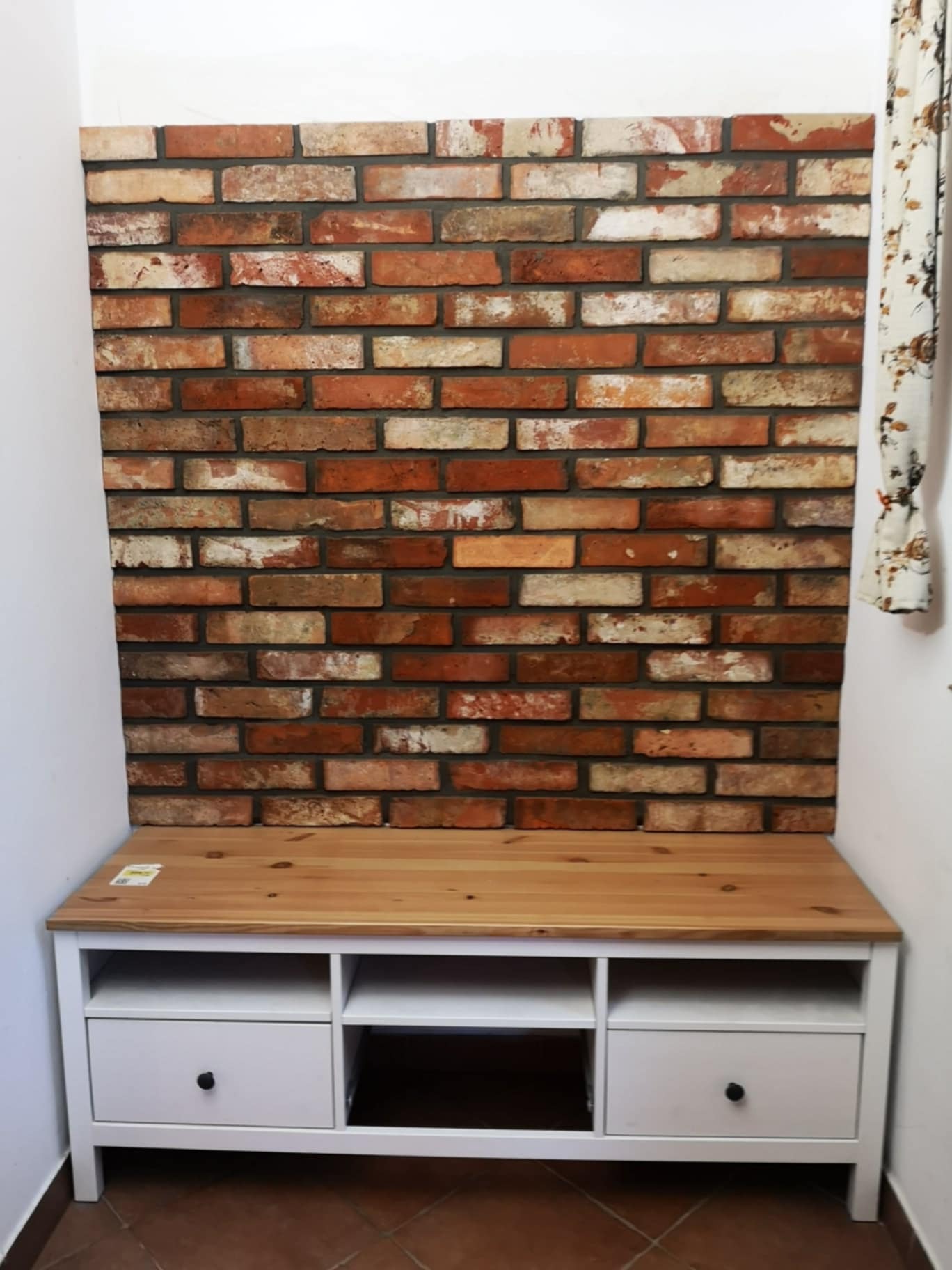Dekoracyjna ściana z cegły klinkierowej za białą szafką RTV z drewnianym blatem, fragment zasłony w tle.
