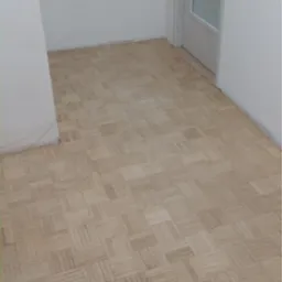 Parkiet ułożony w kwadraty w jasnym pomieszczeniu z białą ścianą i drzwiami z przeszkleniami.