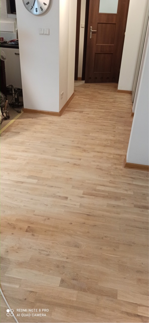 Jasna, odnowiona podłoga parkietowa z wyraźnym rysunkiem drewna, widok na korytarz z brązowymi drzwiami i białą ścianą z zegarem.