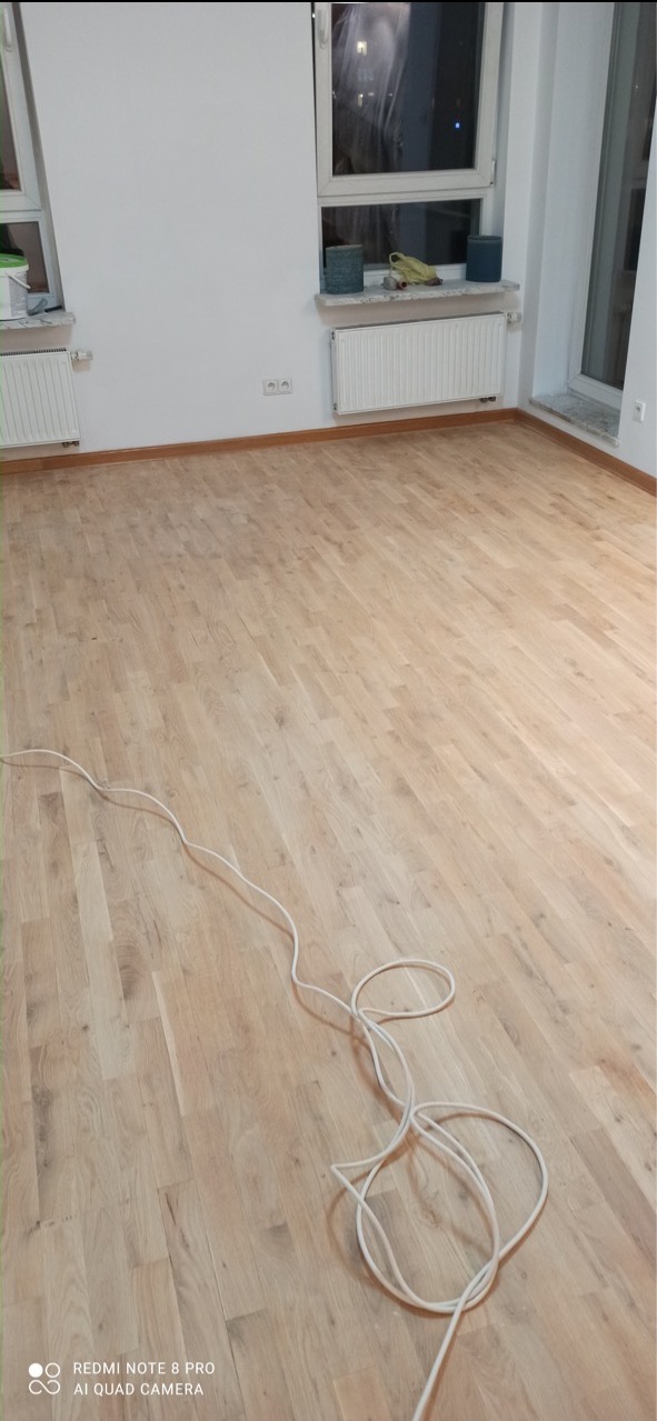 Jasna, odnowiona podłoga parkietowa w pustym pokoju z białymi ścianami, grzejnikami i zwisającym kablem od maszyny do cyklinowania.