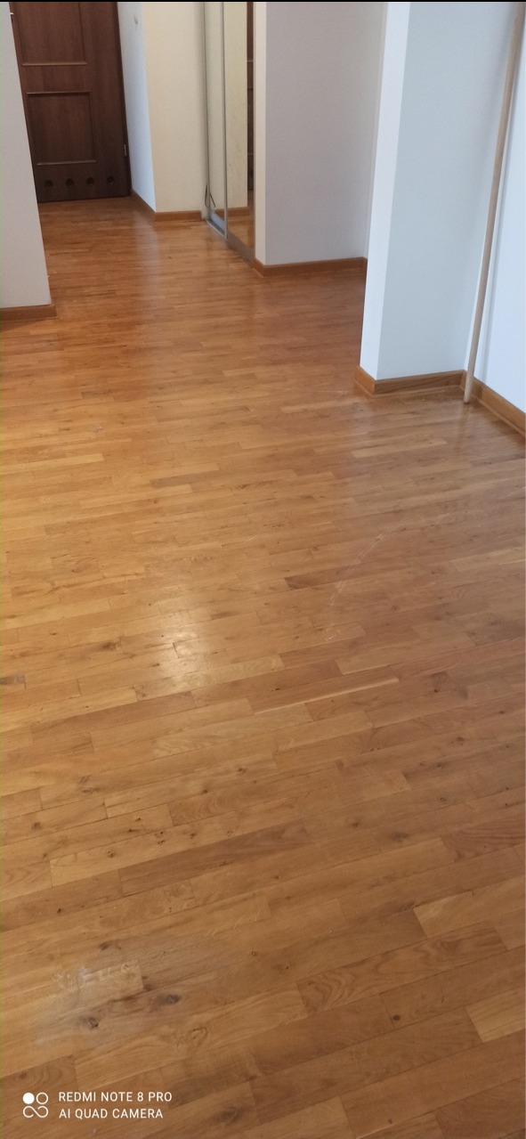 Wypolerowana, jasnobrązowa podłoga parkietowa z widocznymi słojami drewna w pustym pomieszczeniu z białymi ścianami i brązowymi listwami przypodłogowymi, odbijająca światło.