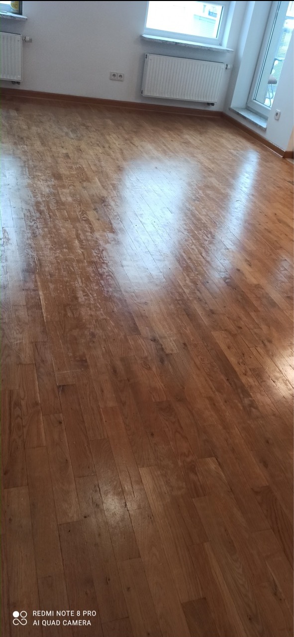 Błyszcząca, odnowiona podłoga drewniana z widocznymi refleksami światła, świeżo po zabiegu renowacji, widoczne grzejniki i okno w tle.