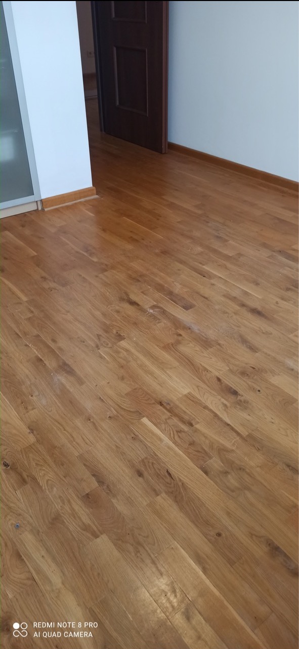 Świeżo wycyklinowana, lakierowana podłoga parkietowa z widocznymi drobnymi pyłkami po szlifowaniu, w tle białe ściany i ciemne drzwi.