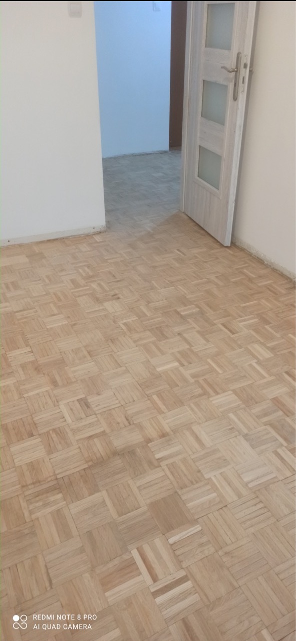 Parkiet ułożony w kwadraty, świeżo po renowacji, widoczne otwarte drzwi do kolejnego pomieszczenia z identyczną podłogą.