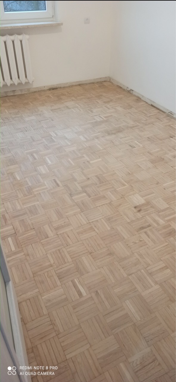 Parkiet ułożony w kwadraty, widoczne ślady po szlifowaniu przy ścianach, pomieszczenie po remoncie, grzejnik żeliwny przy ścianie