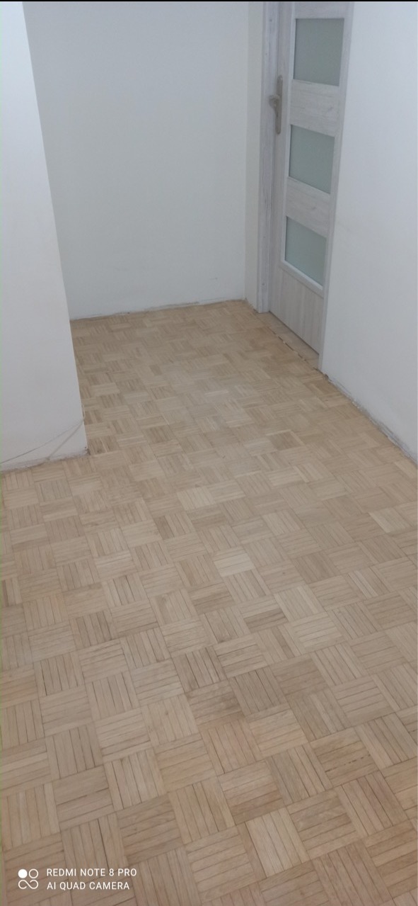 Parkiet ułożony w kwadraty w jasnym pomieszczeniu z białą ścianą i drzwiami z przeszkleniami.