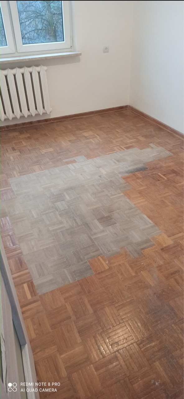 Parkiet po częściowym cyklinowaniu, z widoczną różnicą w kolorze i strukturze drewna, ukazującą efekt odświeżenia podłogi w pokoju z grzejnikiem.