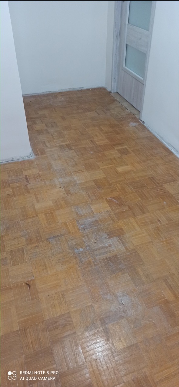 Fragment drewnianej podłogi parkietowej w trakcie renowacji, widoczne ślady szlifowania i pyłu, jasne ściany i białe drzwi w tle.