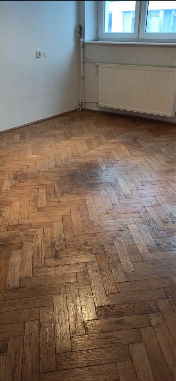 Parkiet ułożony w jodełkę przed cyklinowaniem, widoczne ciemniejsze i jaśniejsze fragmenty, ściana z gniazdkiem elektrycznym, grzejnik pod oknem.