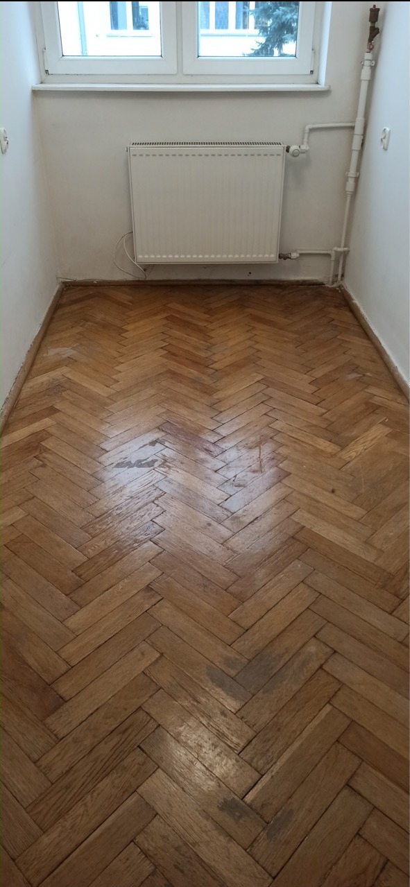 Parkiet ułożony w jodełkę przed cyklinowaniem, widoczne ślady użytkowania i zarysowania, biały grzejnik na ścianie, okno w tle.