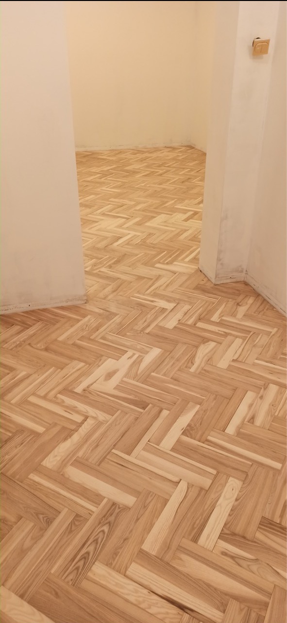 Parkiet ułożony w jodełkę w jasnym pomieszczeniu, widoczne niedoskonałości wykończenia przy ścianach.