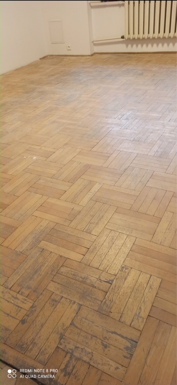 Parkiet ułożony w jodełkę przed renowacją, widoczne ślady zużycia i zarysowania, jasne ściany i grzejnik w tle.