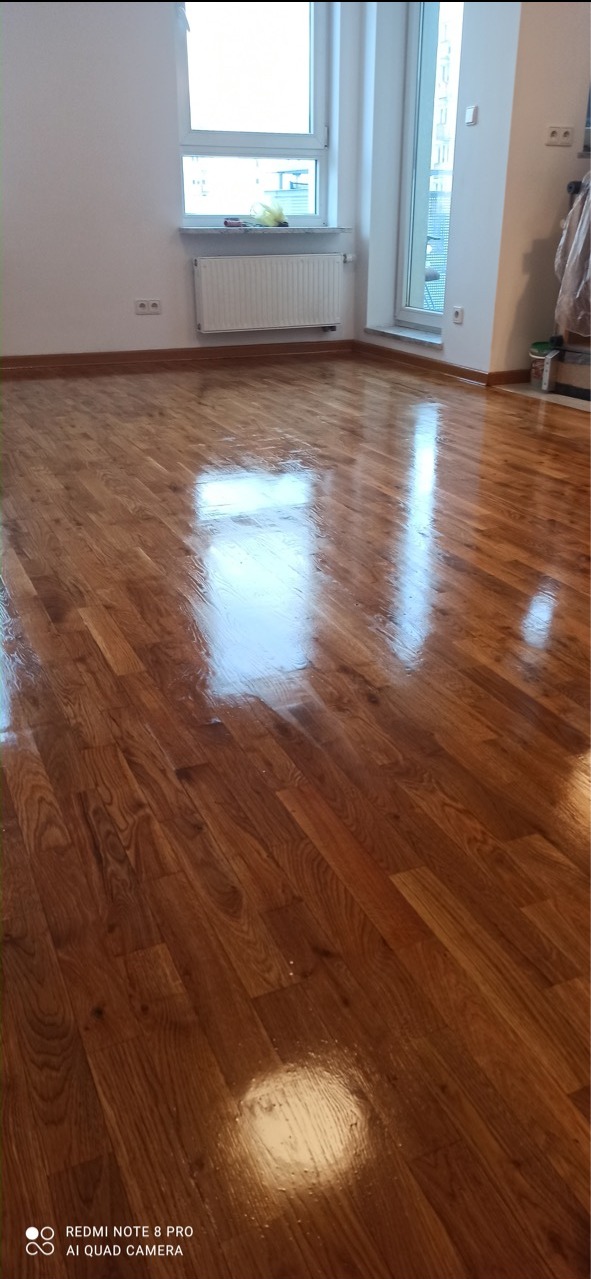 Błyszcząca, świeżo polakierowana podłoga parkietowa w pustym pokoju z białymi ścianami, grzejnikiem i oknem balkonowym.