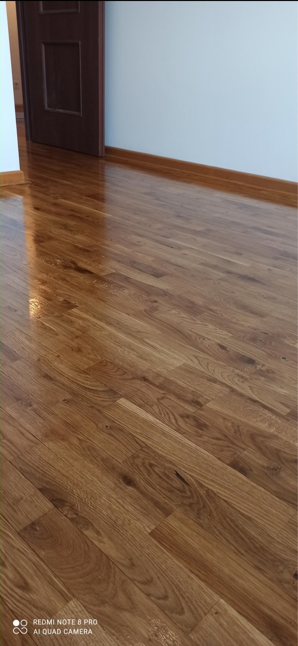 Błyszcząca, świeżo polakierowana podłoga parkietowa z widoczną listwą przypodłogową i fragmentem otwartych, ciemnych drzwi.