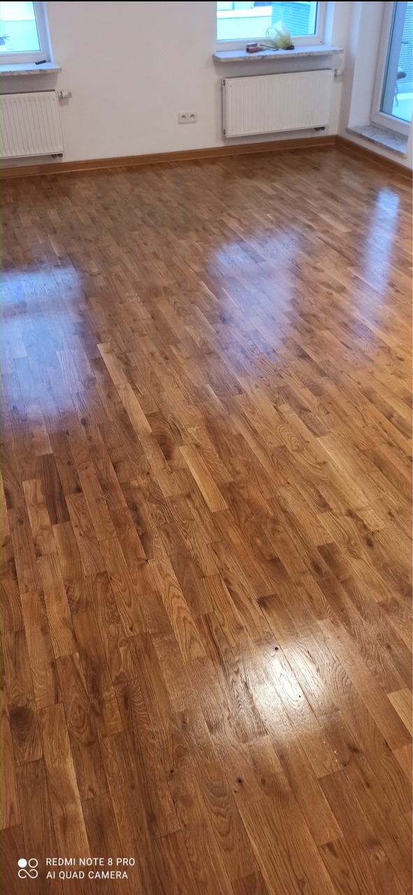 Błyszcząca, odnowiona podłoga parkietowa w pustym pokoju z białymi ścianami, grzejnikami i oknami.