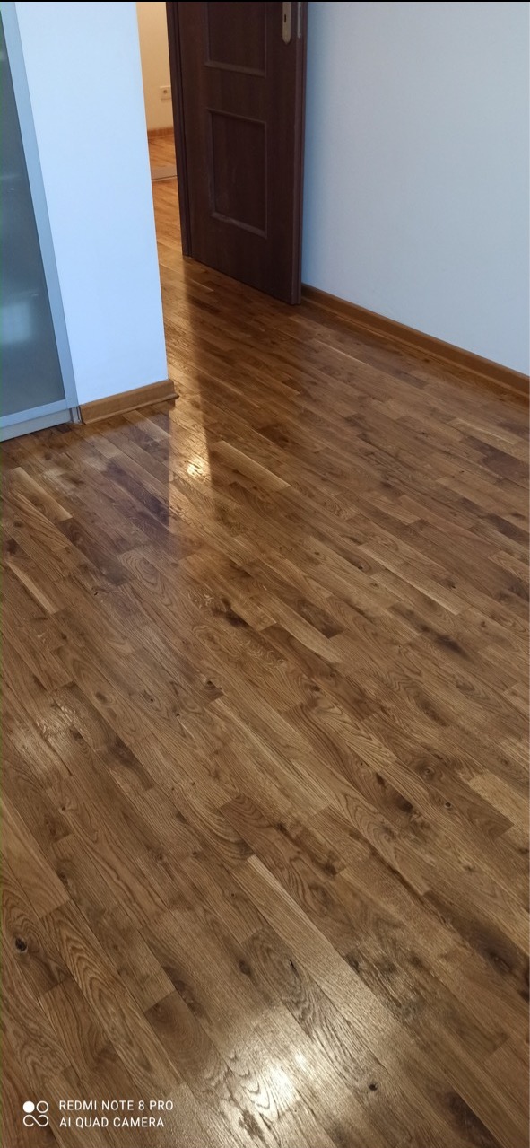 Parkiet dębowy po renowacji, widoczny połysk lakieru, fragment otwartych drzwi i biała ściana.