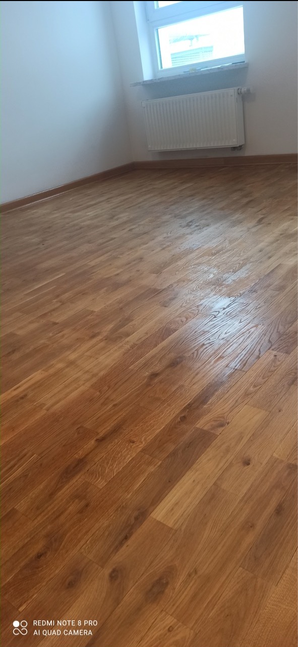 Wycyklinowana podłoga z parkietu w odcieniach brązu, widoczny fragment ściany i grzejnik pod oknem.