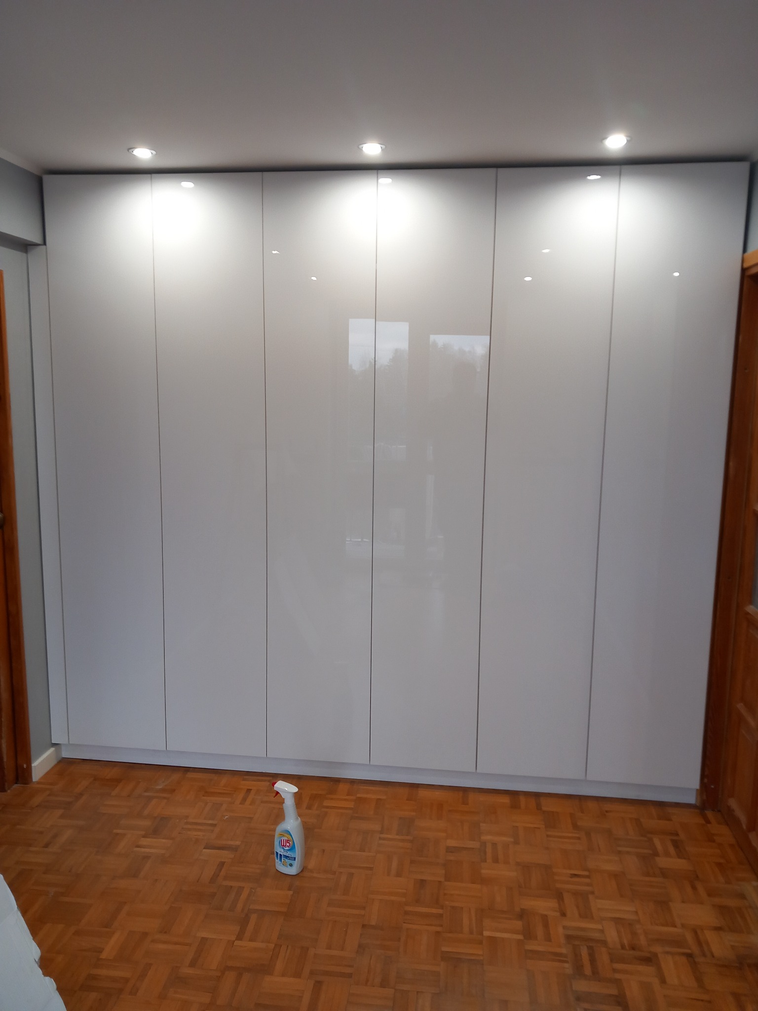 Błyszcząca, biała zabudowa szafy wnękowej od podłogi do sufitu z oświetleniem punktowym, widoczna butelka środka czyszczącego na parkiecie.