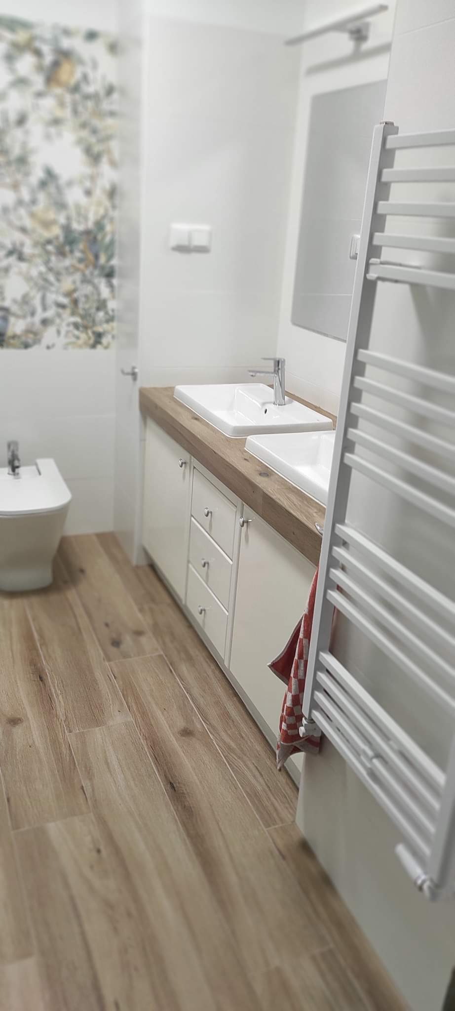 Nowoczesna łazienka z dwoma umywalkami na drewnianym blacie, białe szafki z szufladami i chromowanymi uchwytami, drewnopodobna podłoga, grzejnik drabinkowy i bidet, akcent w postaci ręcznika...
