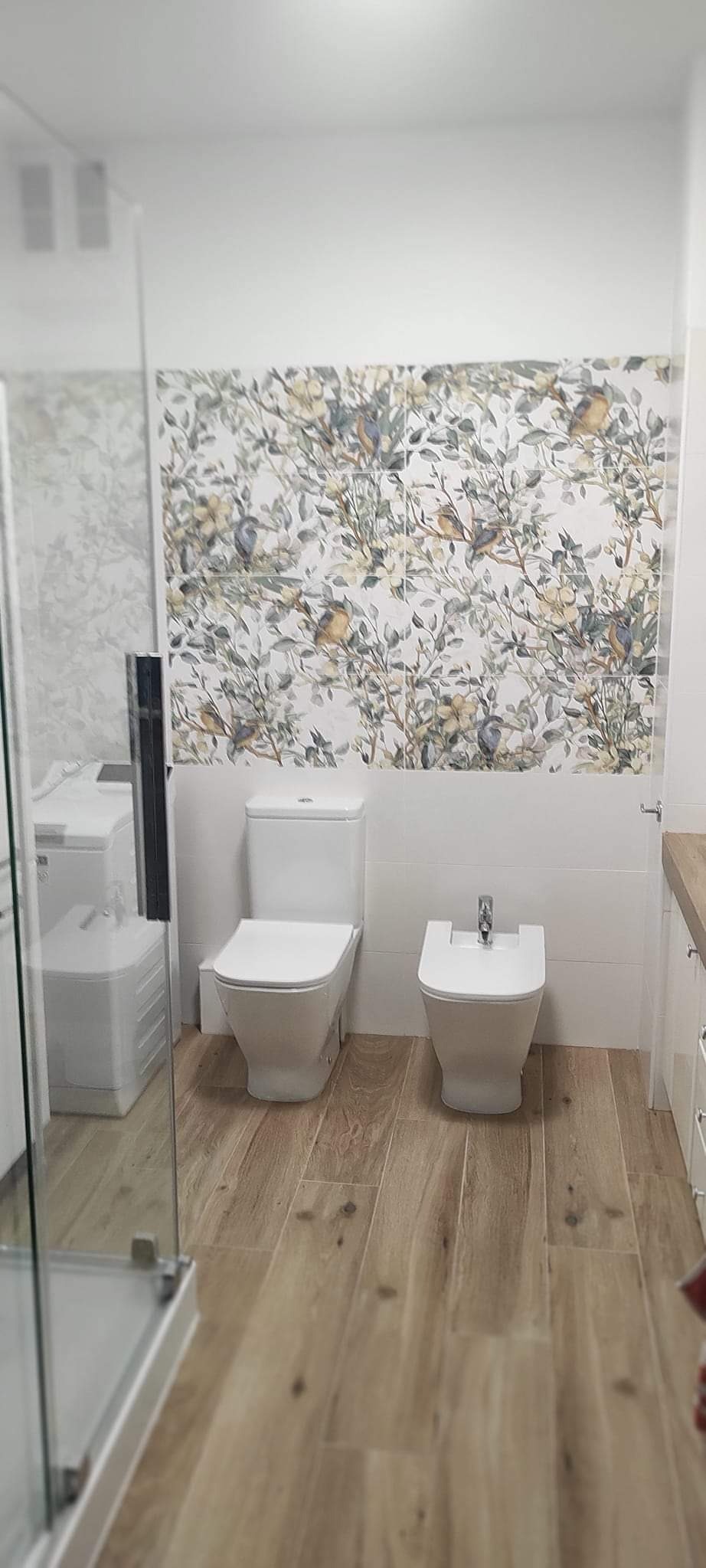 Wykończona łazienka z drewnopodobną podłogą, białą armaturą (toaleta i bidet) oraz dekoracyjną ścianą z płytek w motywy roślinne i ptasie.