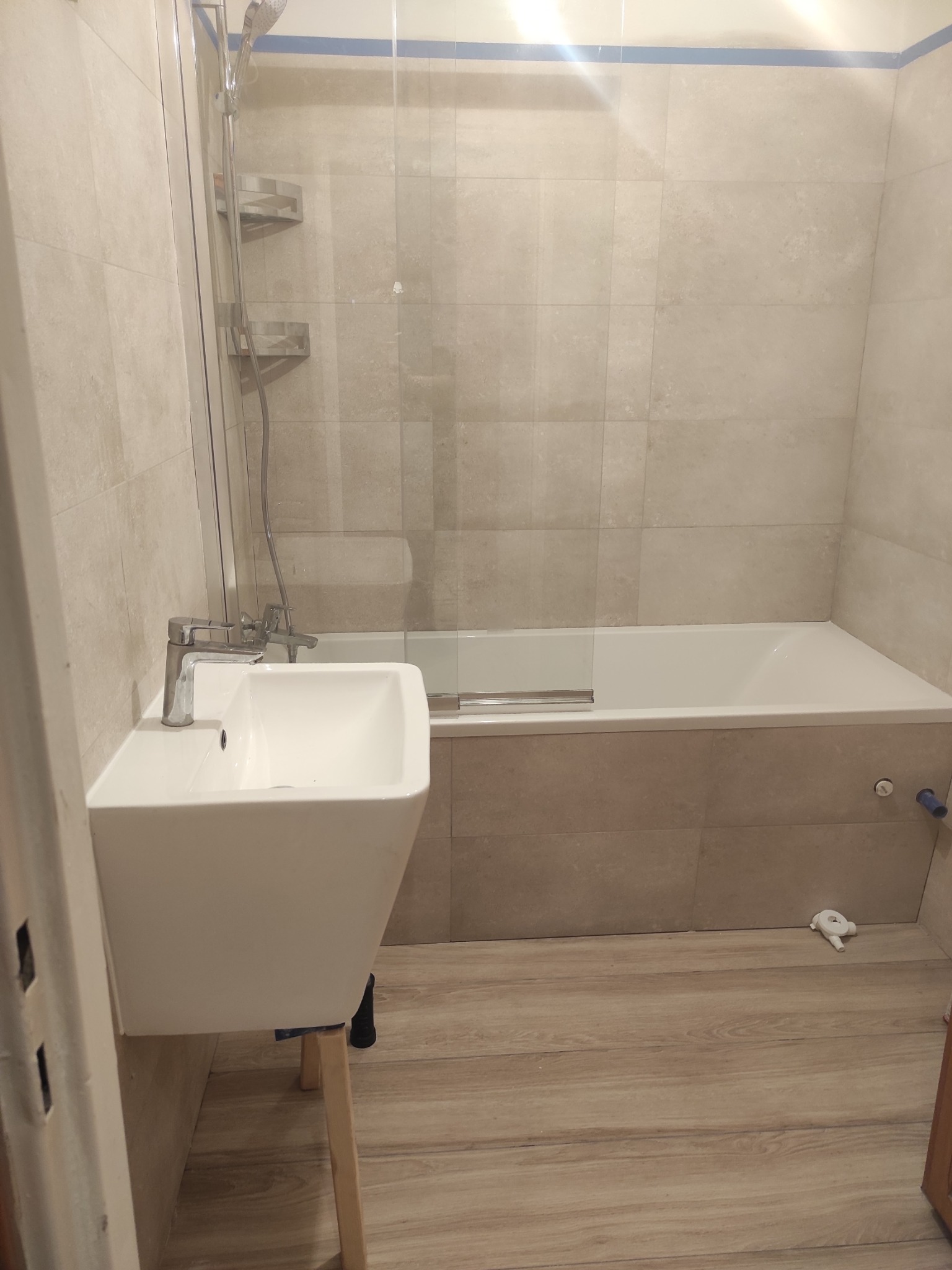 Ukończona łazienka w beżowych odcieniach: prostokątna umywalka na drewnianych nóżkach, przeszklona kabina prysznicowa nad wanną, płytki imitujące drewno na podłodze, niebieska taśma malarska przy...